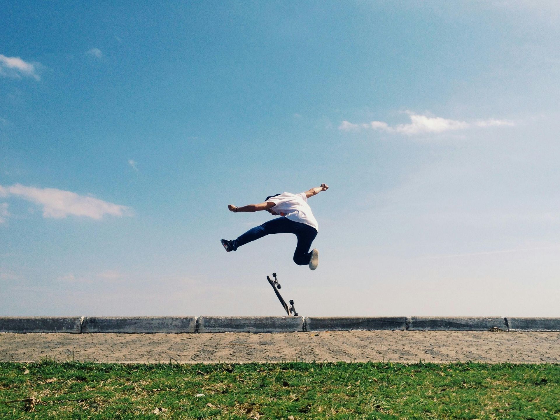 Person mid-jump over a skateboard on a grassy field under a blue sky