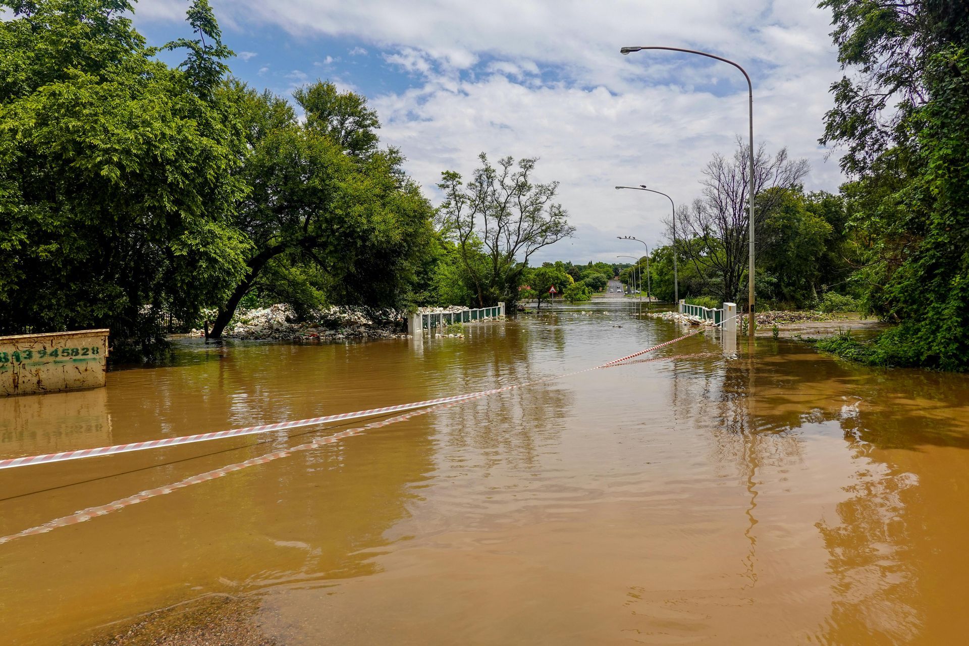 Flooded street lined with trees and lampposts under a partly cloudy sky