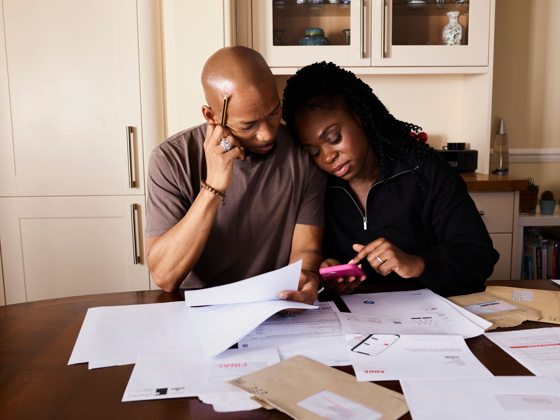 Two people review paperwork at a kitchen table, one holding a pink phone amid scattered documents.