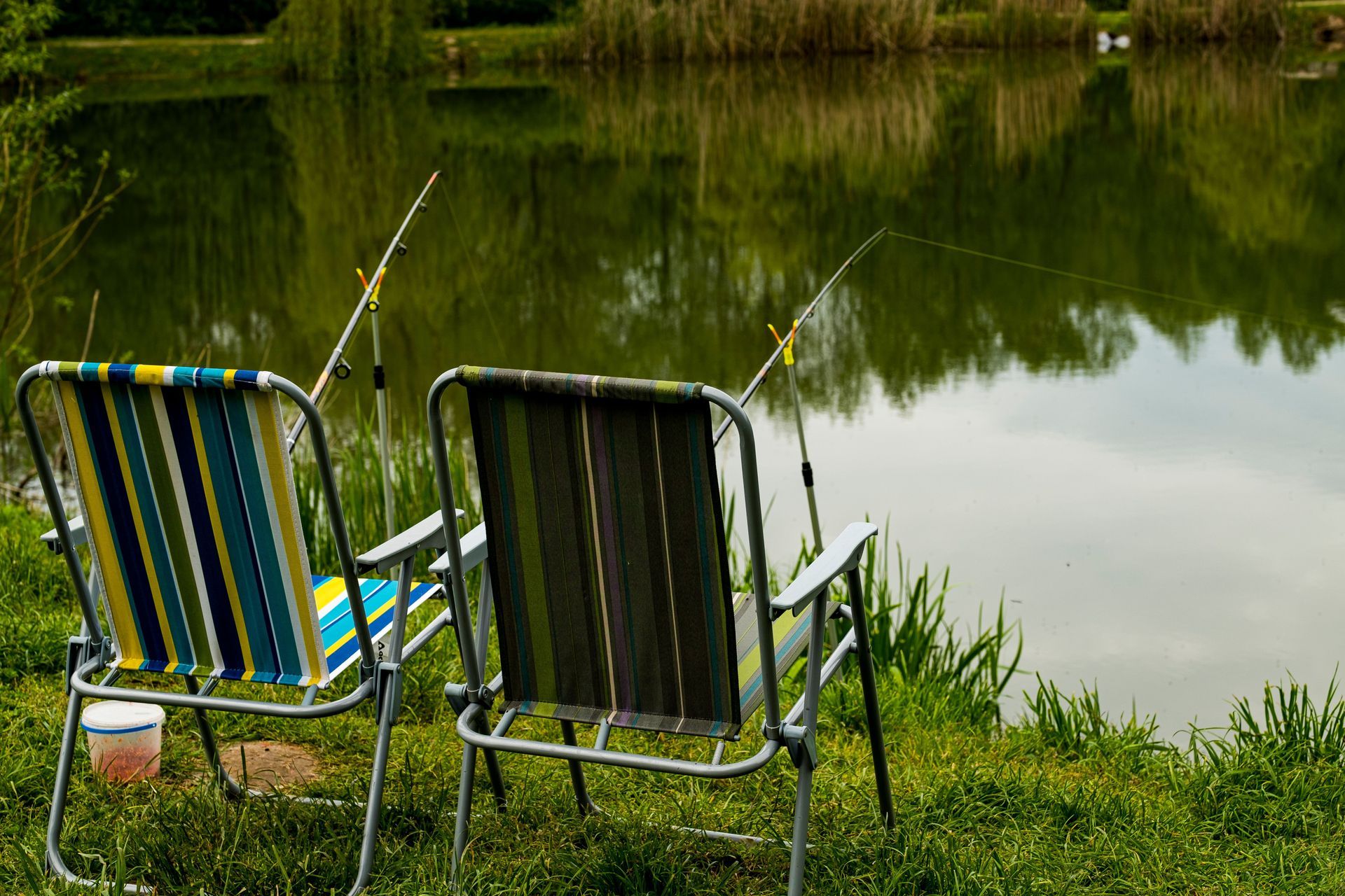 Two folding lawn chairs and fishing rods on a grassy pond bank, where weekend guests gather by water