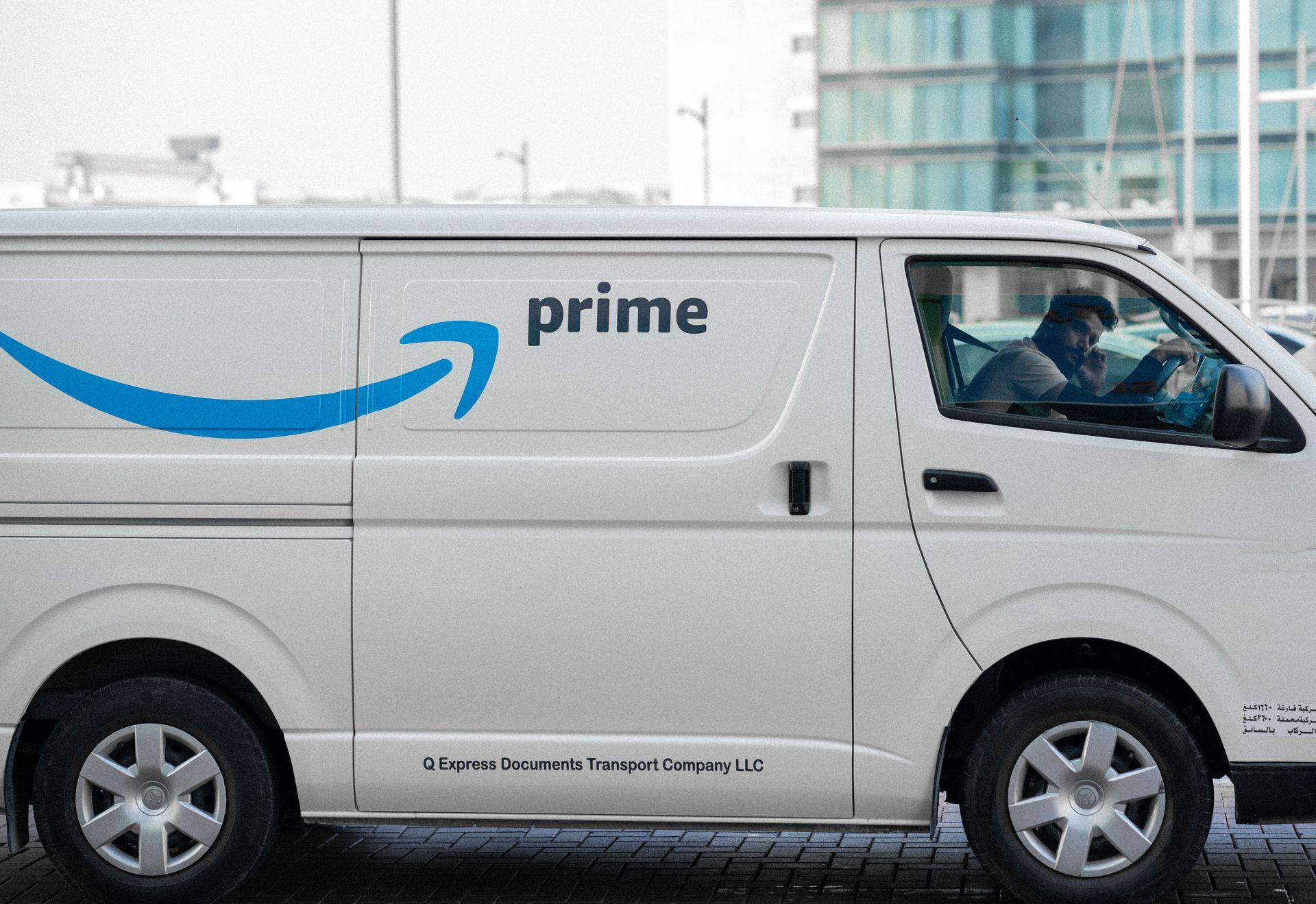 White Amazon Prime delivery van parked on a street, side view with driver visible.