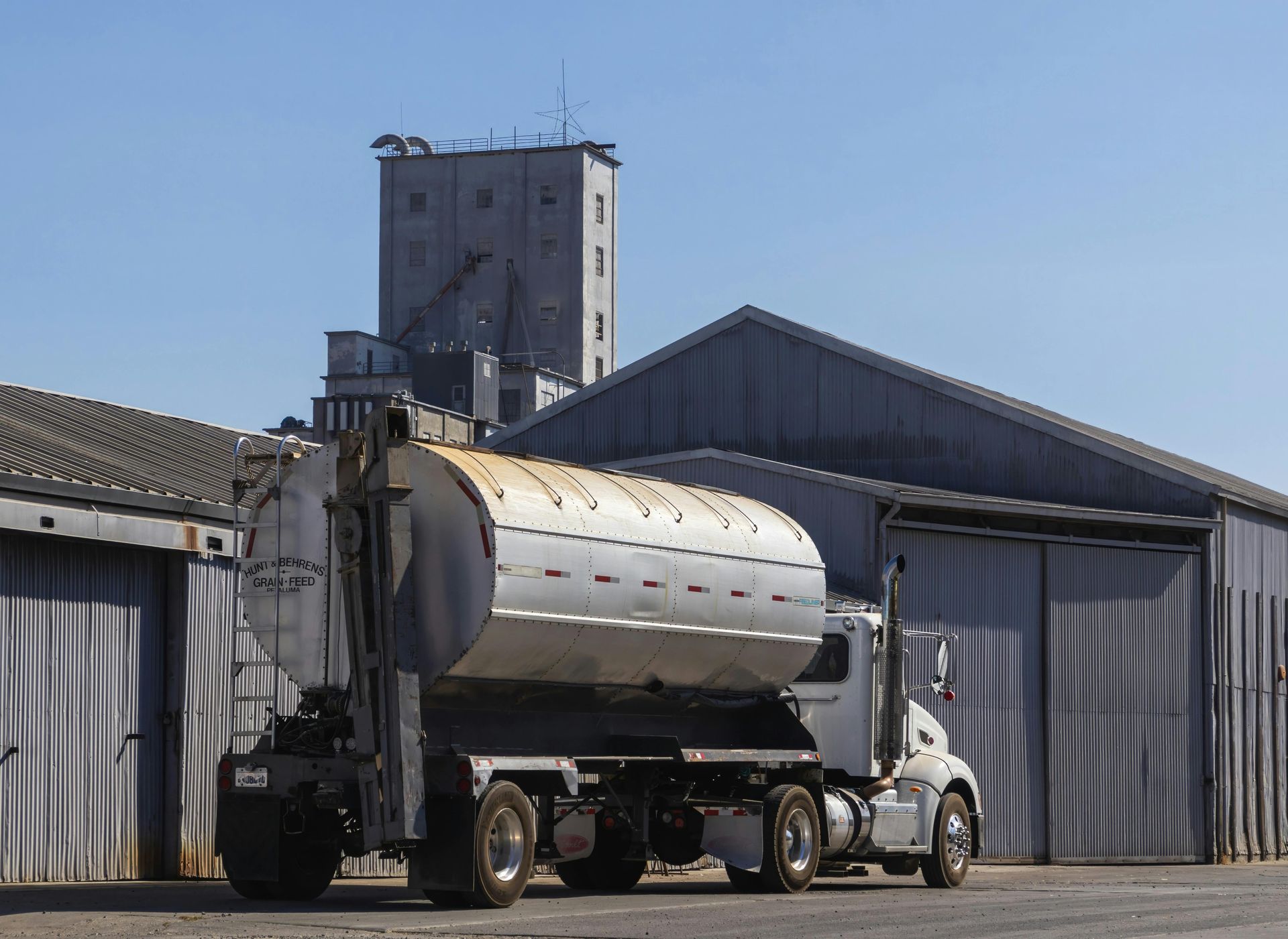 Silver tanker truck parked beside a gray industrial warehouse under a clear sky