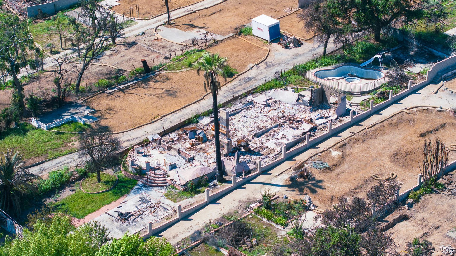 Damaged roadside area with debris, a parked car, and dry trees near a small round fountain
