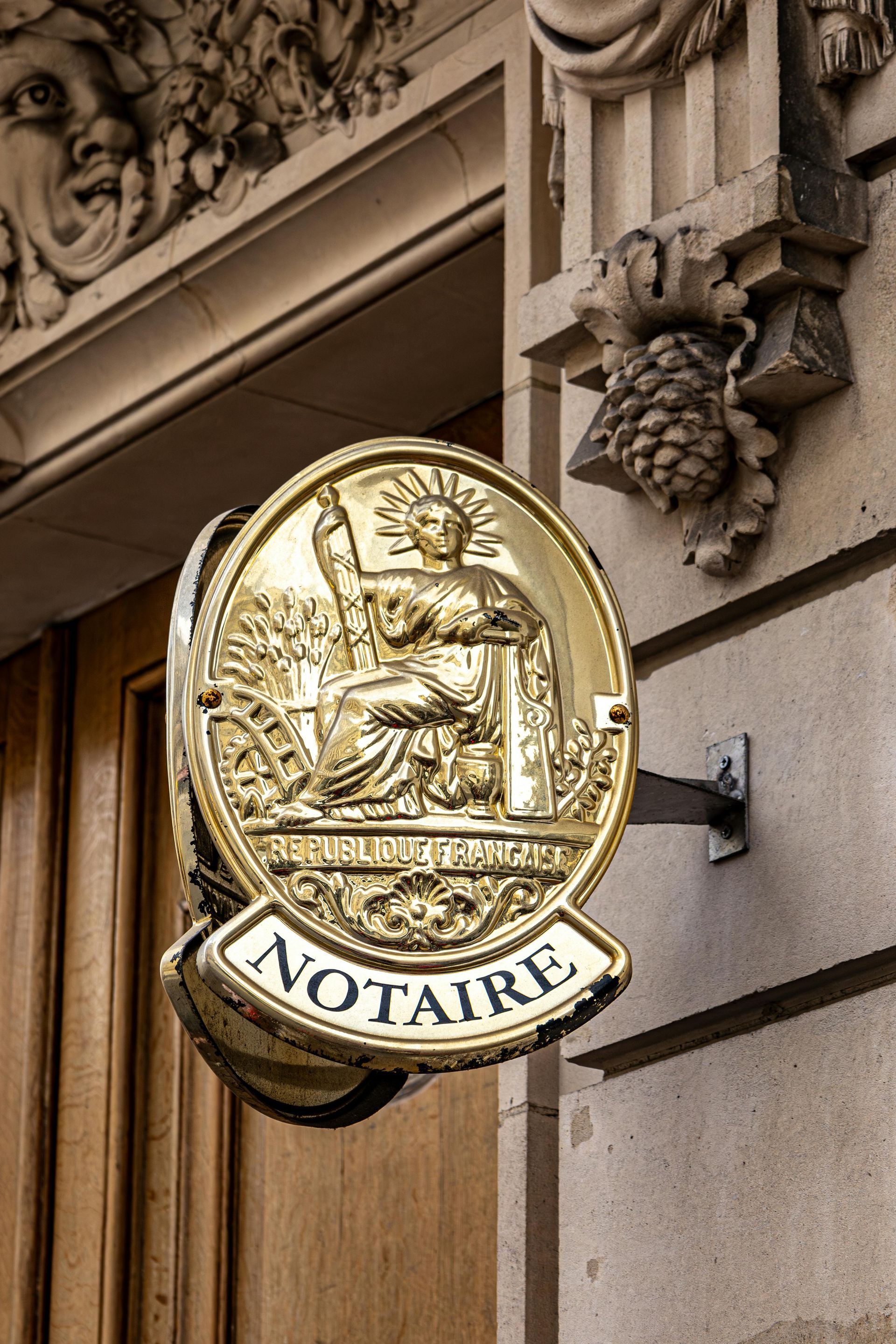 Gold notary sign on a stone building facade with “NOTAIRE” text and grape detail
