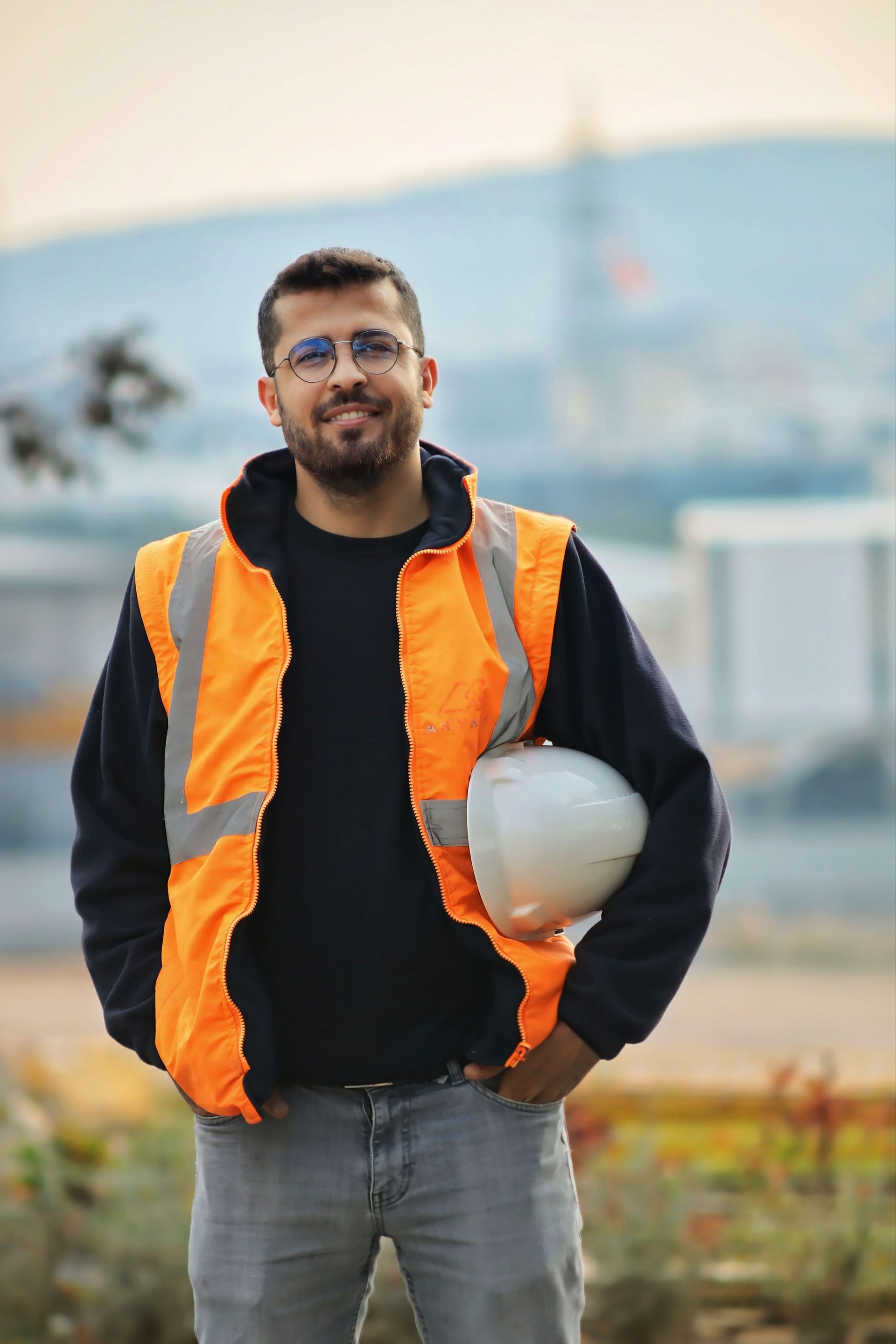 Man in orange safety vest holding a hard hat outdoors near a construction site