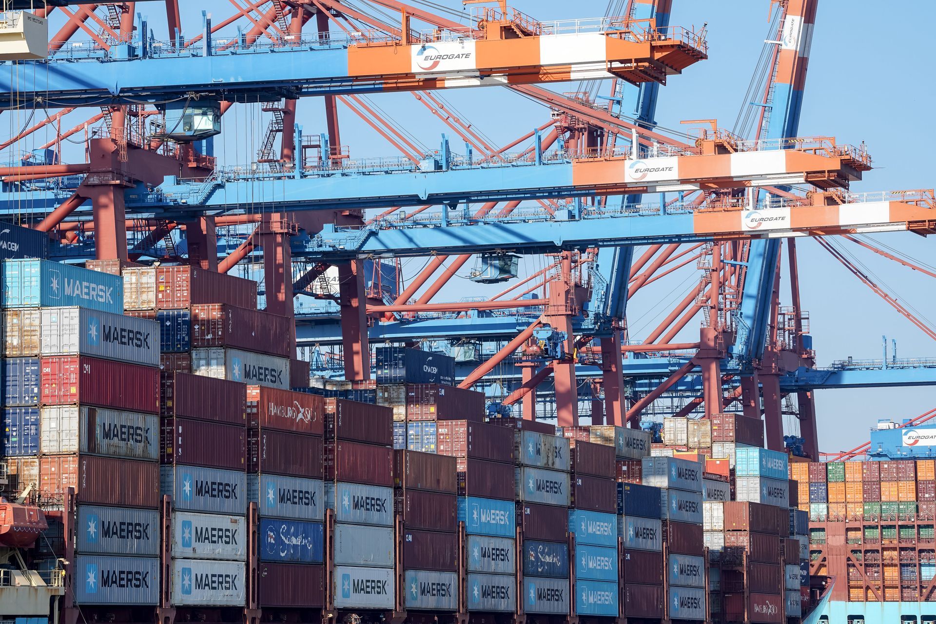Stacked shipping containers in a busy port with orange cranes against a blue sky