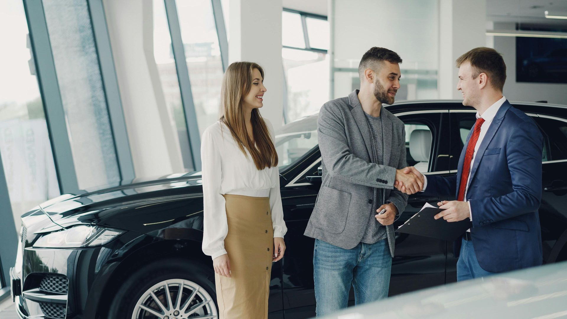 Three people shaking hands beside a black car in a bright showroom