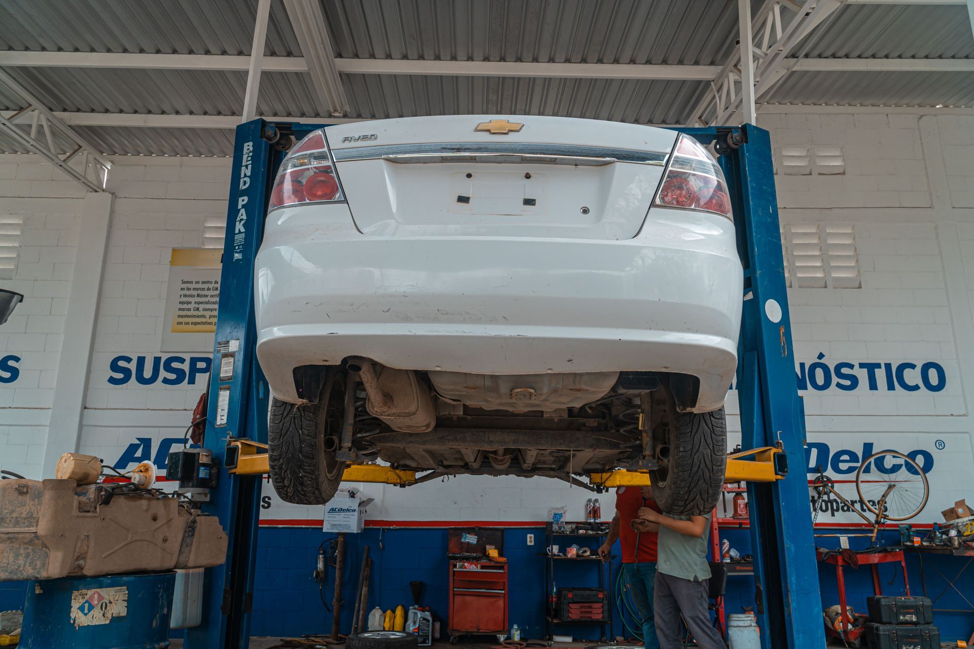White sedan lifted on a blue two-post hoist inside an auto repair shop while a technician works underneath