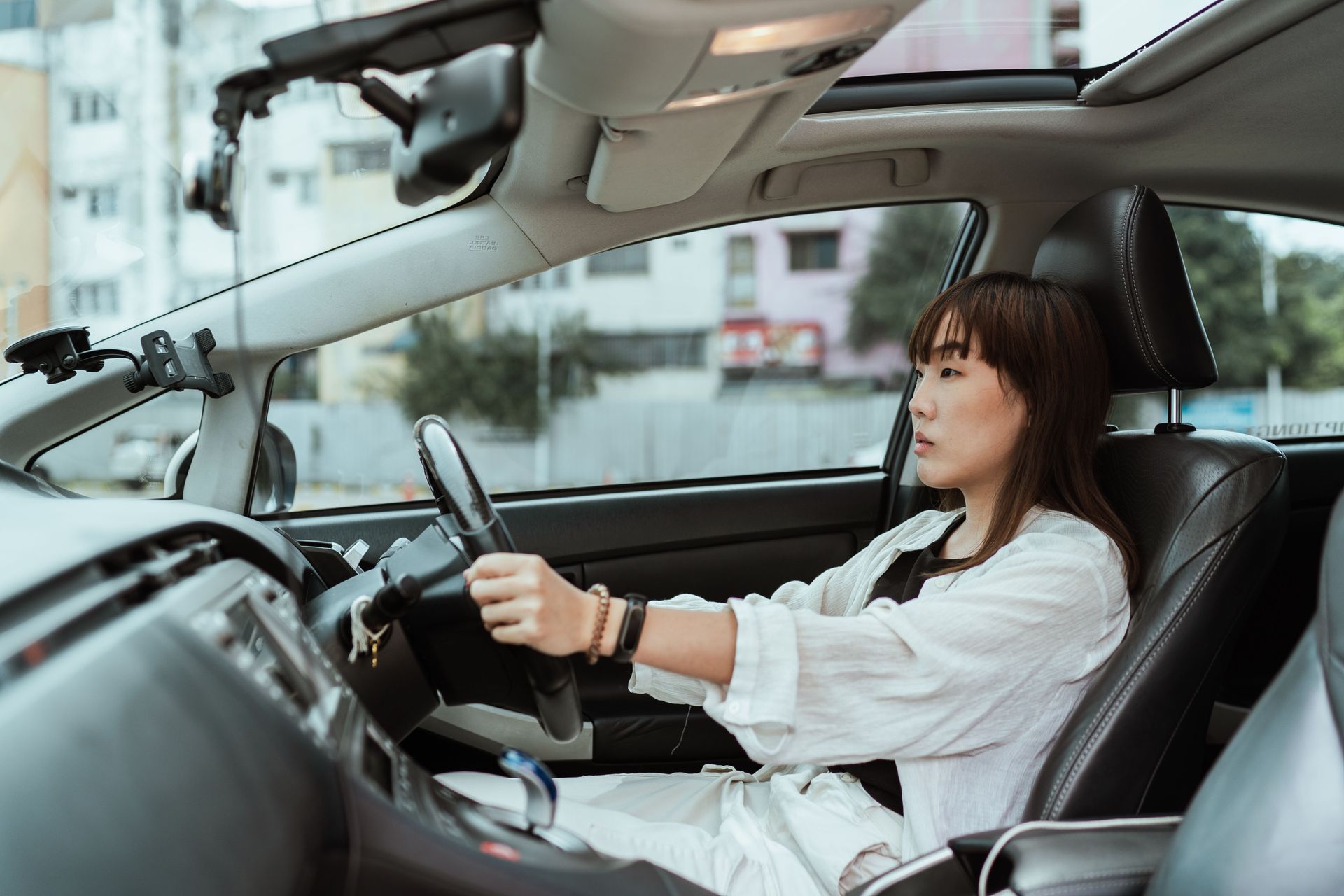 A woman with both hands on the steering wheel drives her car, seen from the passenger seat on a city street.
