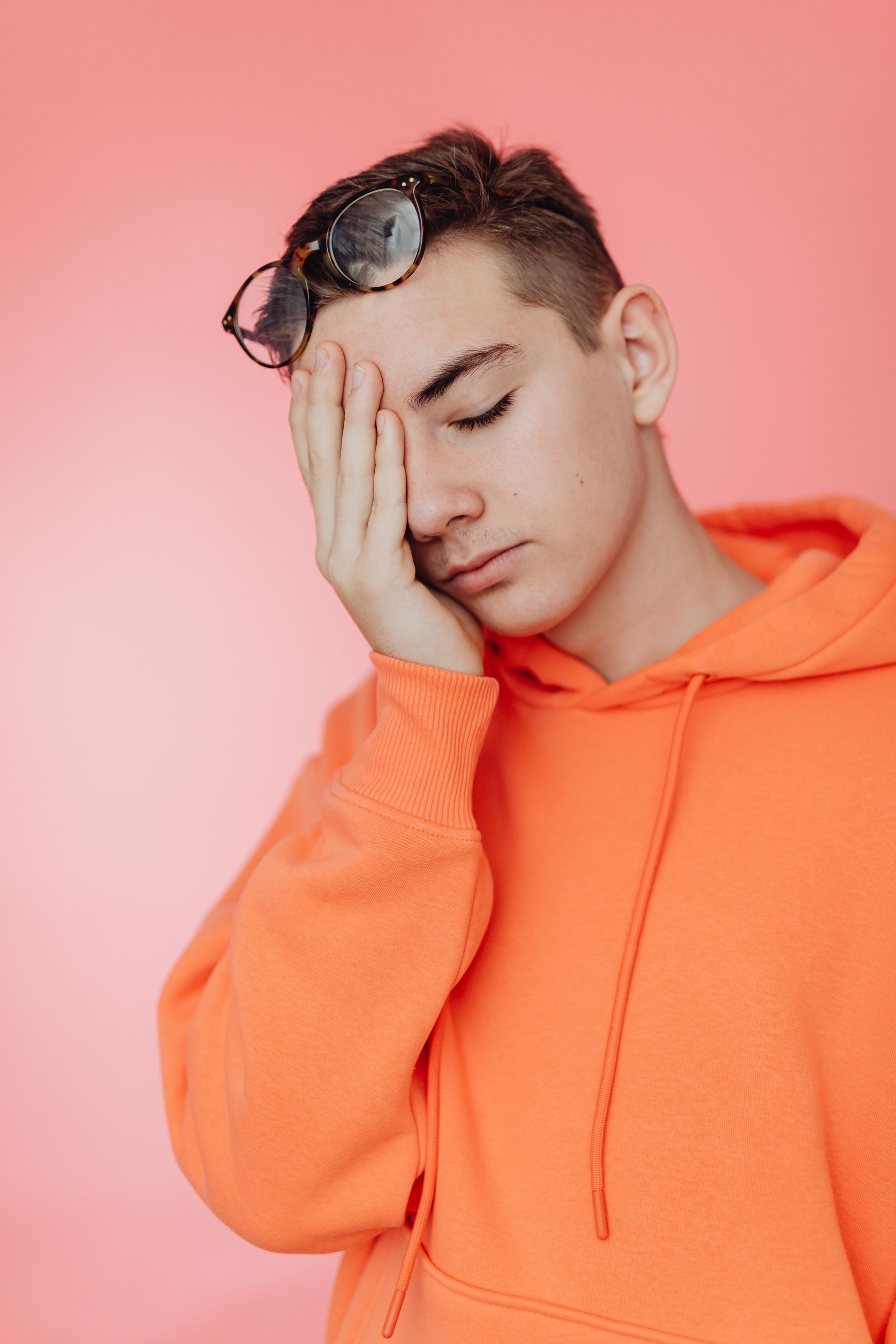 Person in an orange hoodie covering one eye with a hand against a pink background