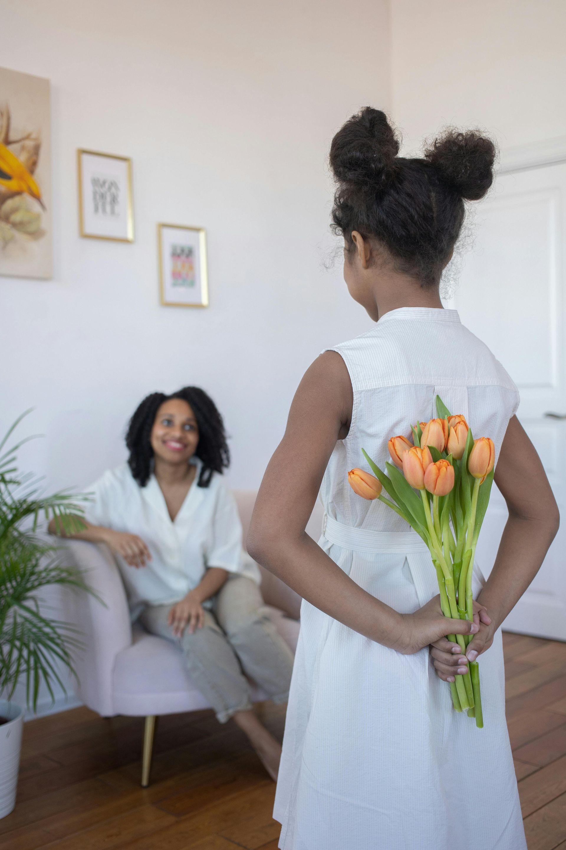 Woman in white holds orange tulips behind her back while facing a seated person in a bright room.