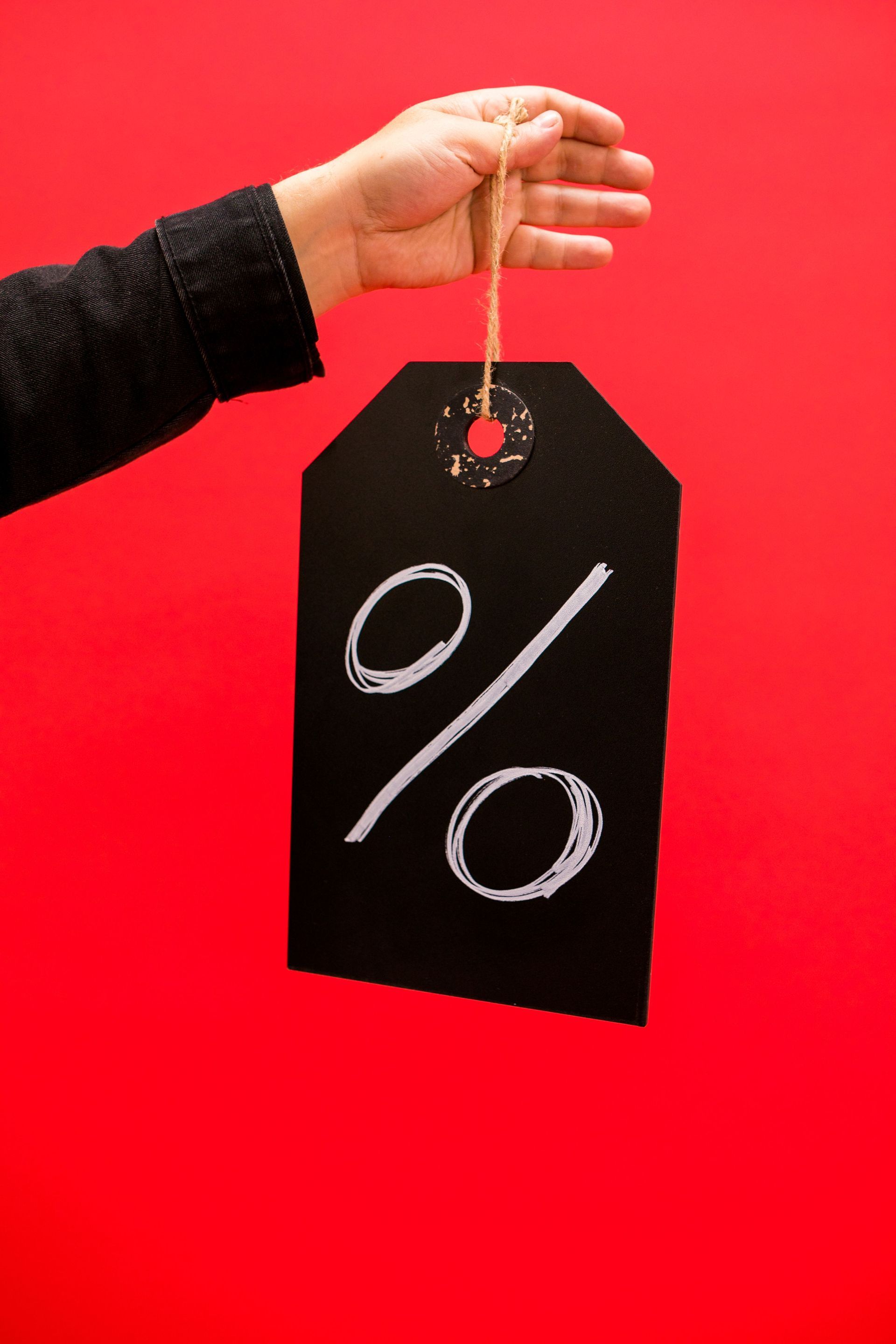 Hand holding a black sale tag with a white percent sign on a red background