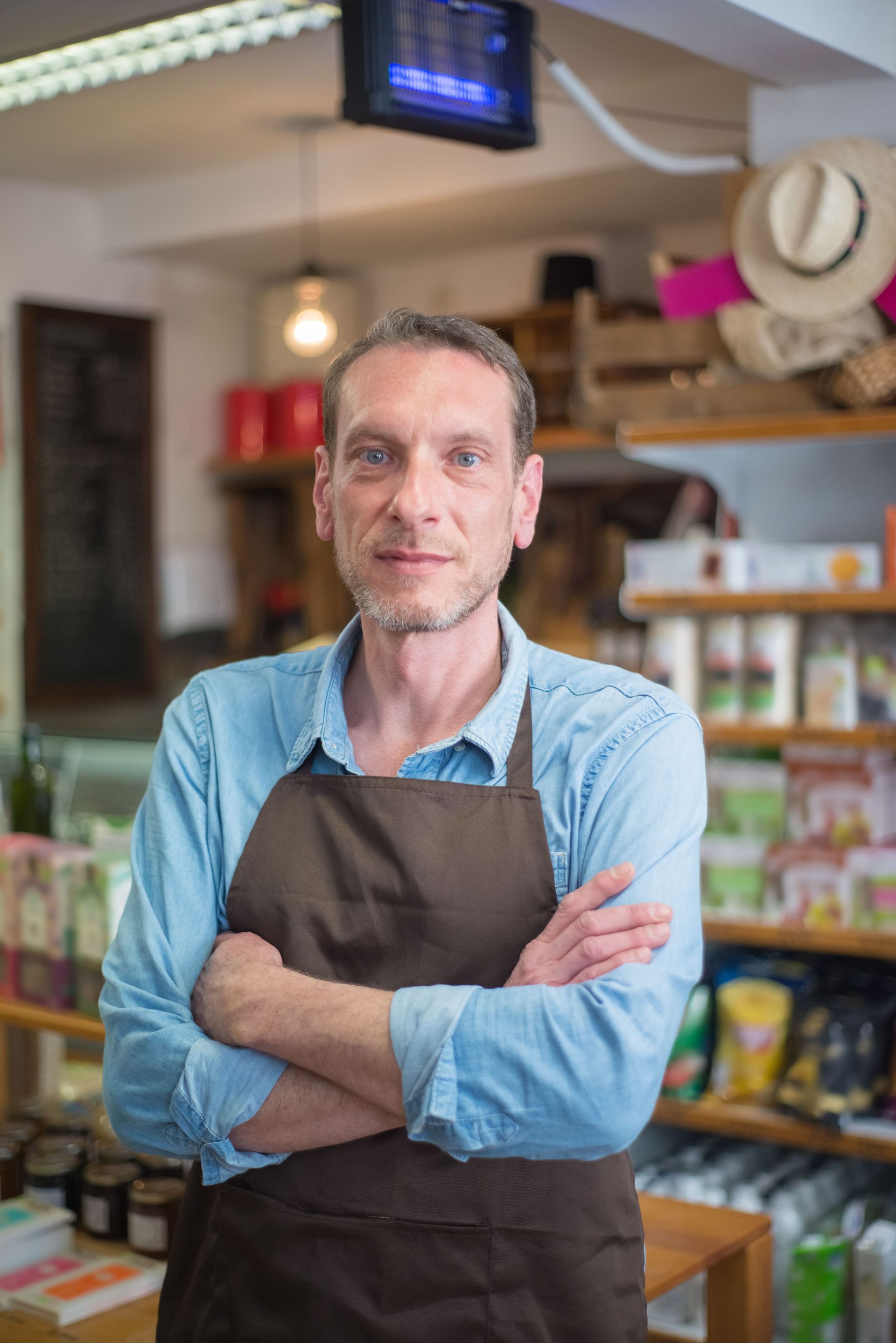 Shopkeeper in a brown apron stands with arms crossed in a small store.