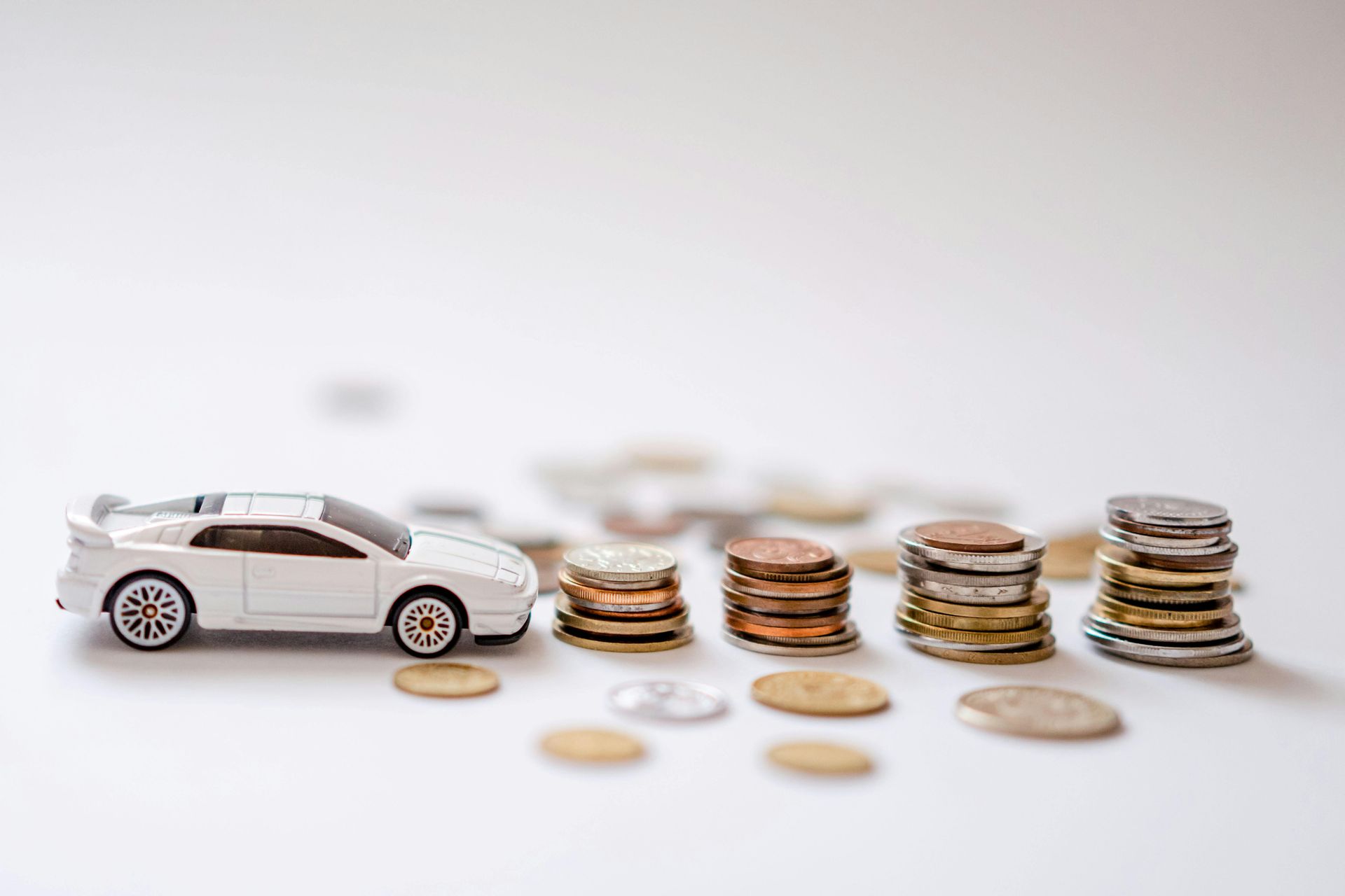 White toy car beside stacks of coins on a white surface, symbolizing car savings or cost.