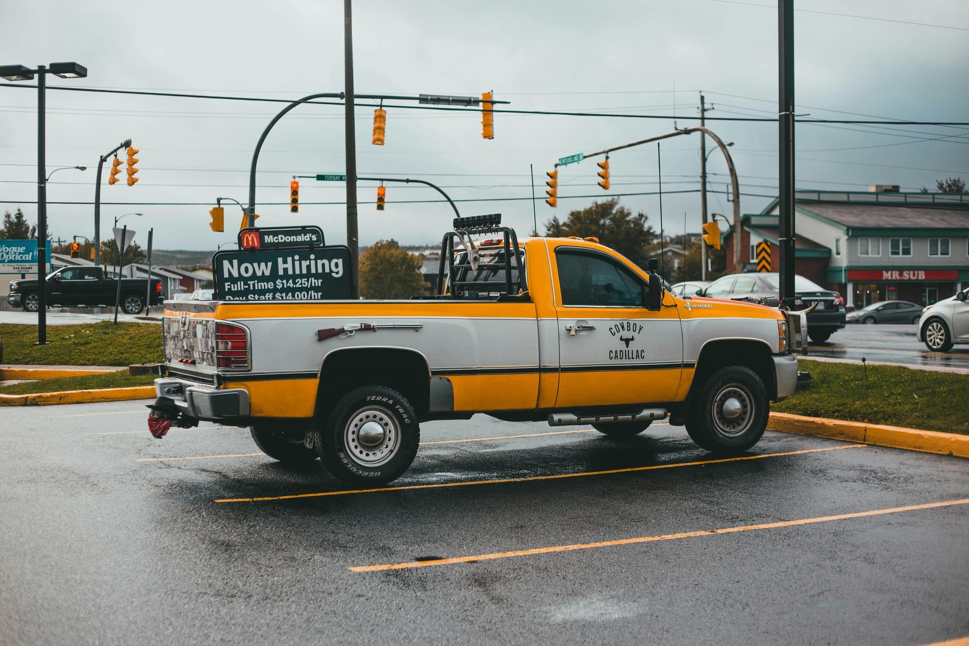 Yellow-and-white tow truck with “Now Hiring” sign driving in a rainy city intersection