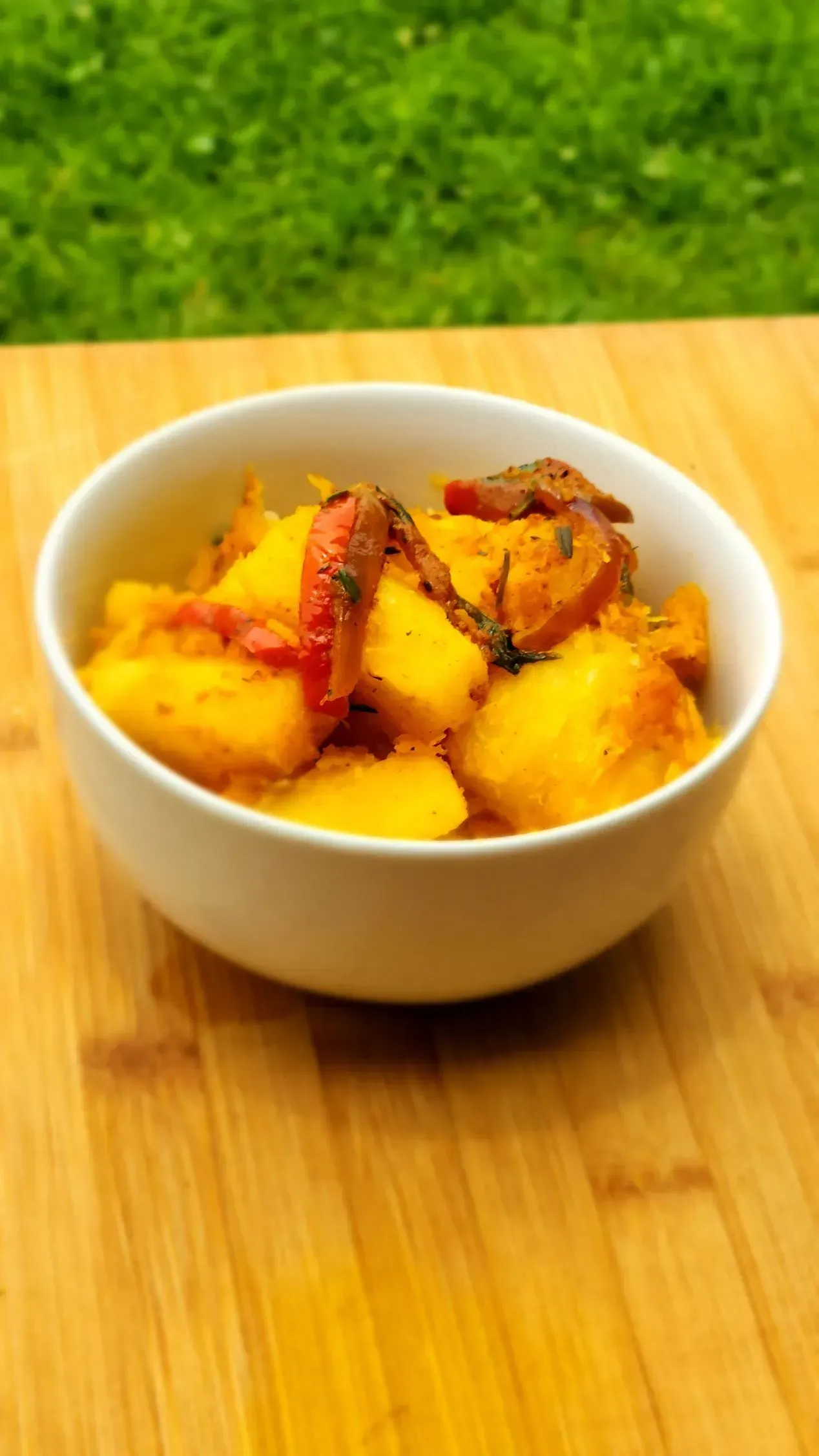 Bowl of yellow food, possibly potatoes or squash, with red garnish, on a wooden board against green grass.