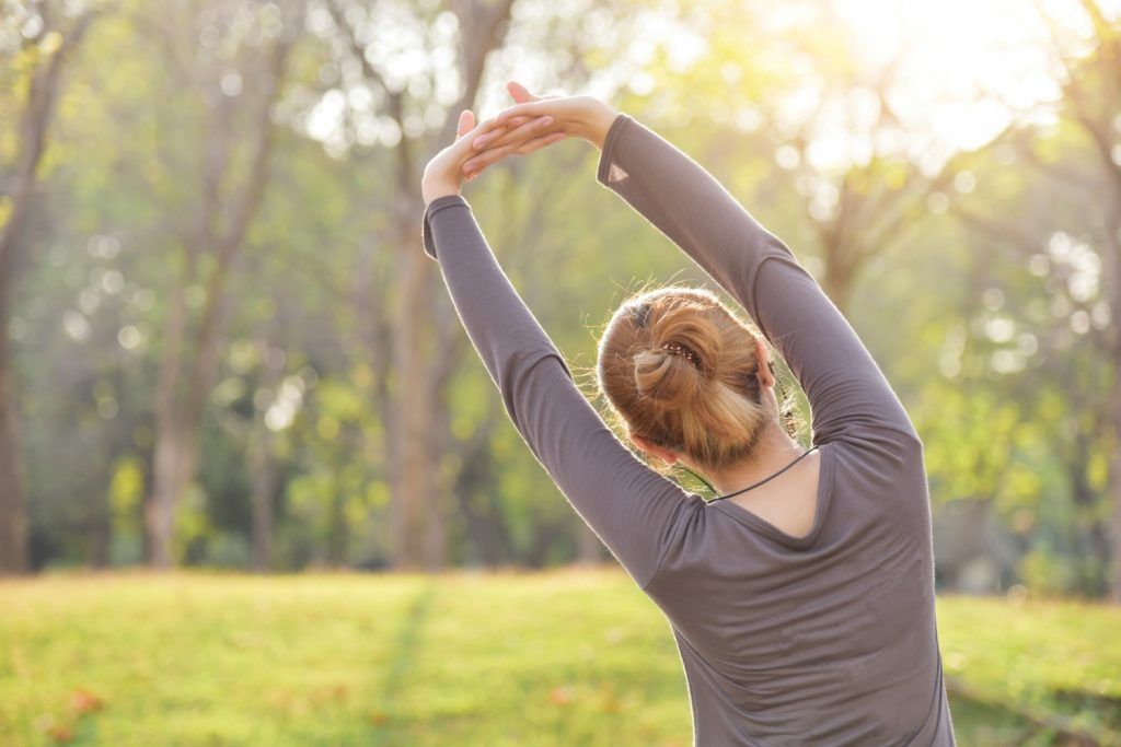 Person stretching outdoors in a sunlit park with arms raised overhead
