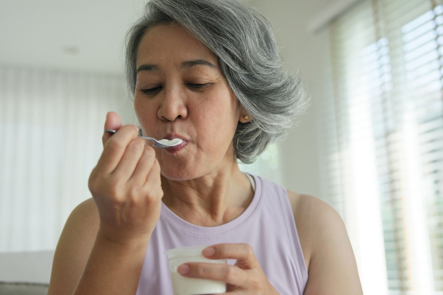 Woman eating yogurt with a spoon in a bright room with window blinds