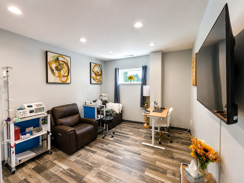 Small medical treatment room with exam chair, stool, cabinet, and black recliner on gray flooring.