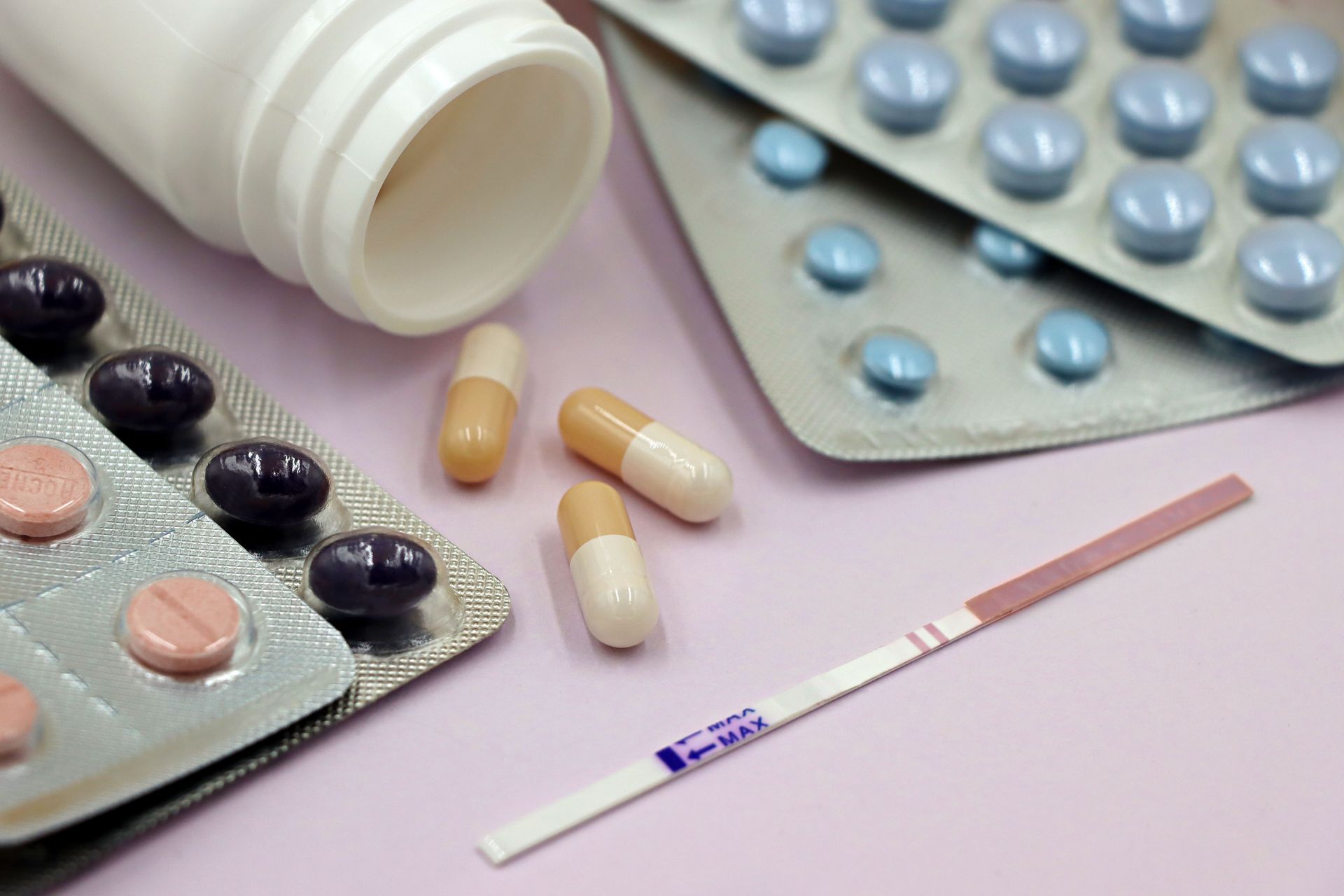 Assorted pills and capsules spilling from a bottle with blister packs and a pregnancy test on a pink surface