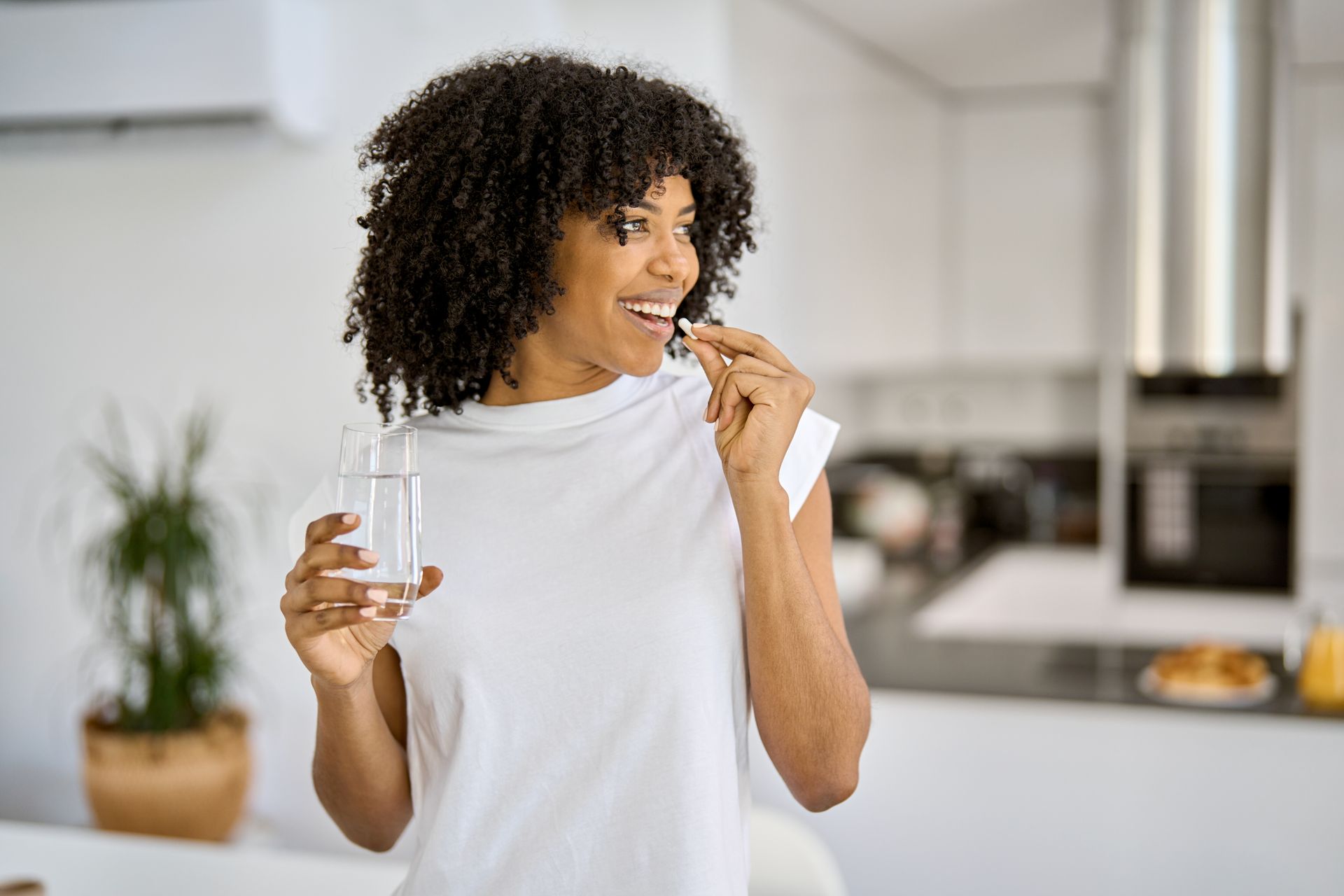 Person holding a pill and water in a bright kitchen, smiling.