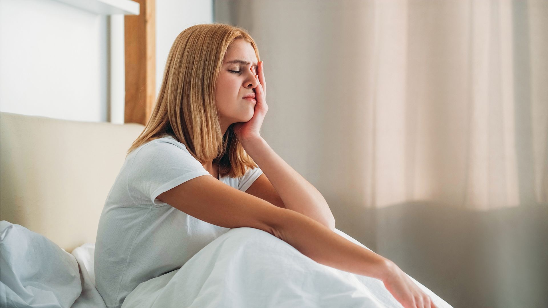 Person sitting on a bed by a window, resting a hand on their chin and looking out thoughtfully.