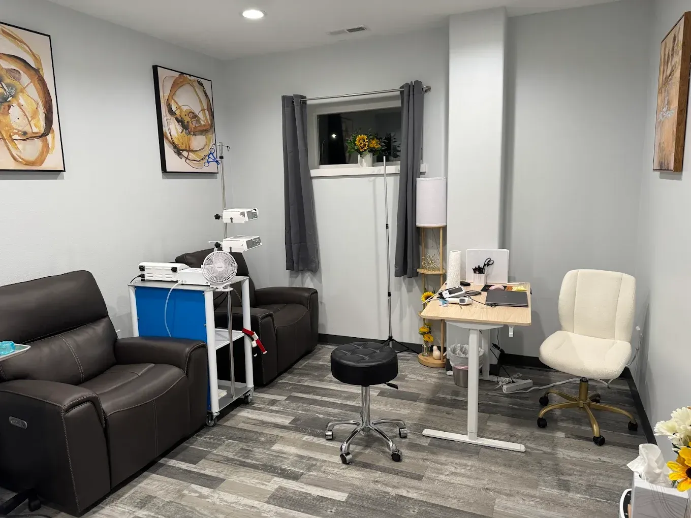Small medical treatment room with exam chair, stool, cabinet, and black recliner on gray flooring.