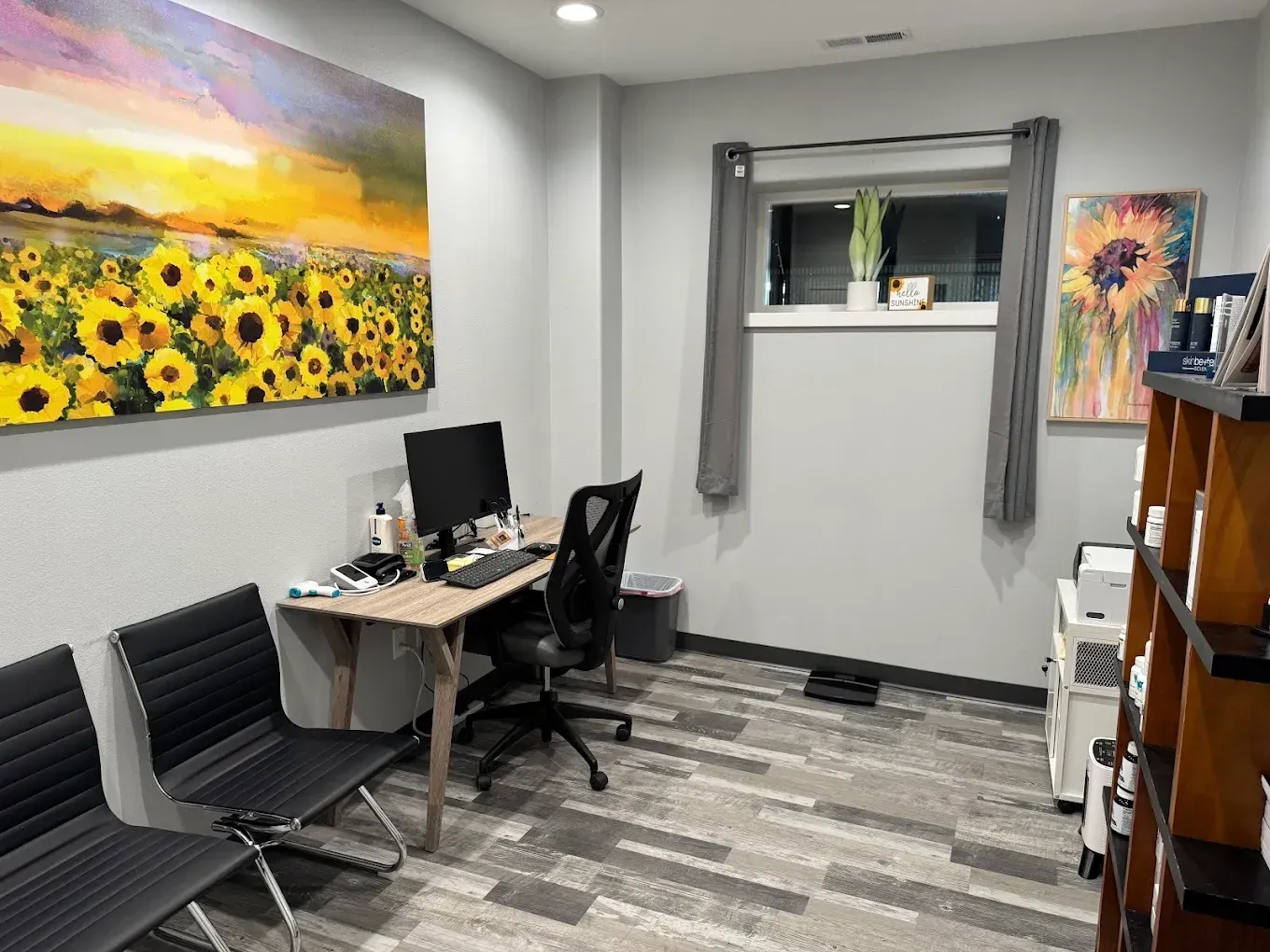 Small office with desk, computer, chairs, gray walls, sunflower painting, and a window with curtains.