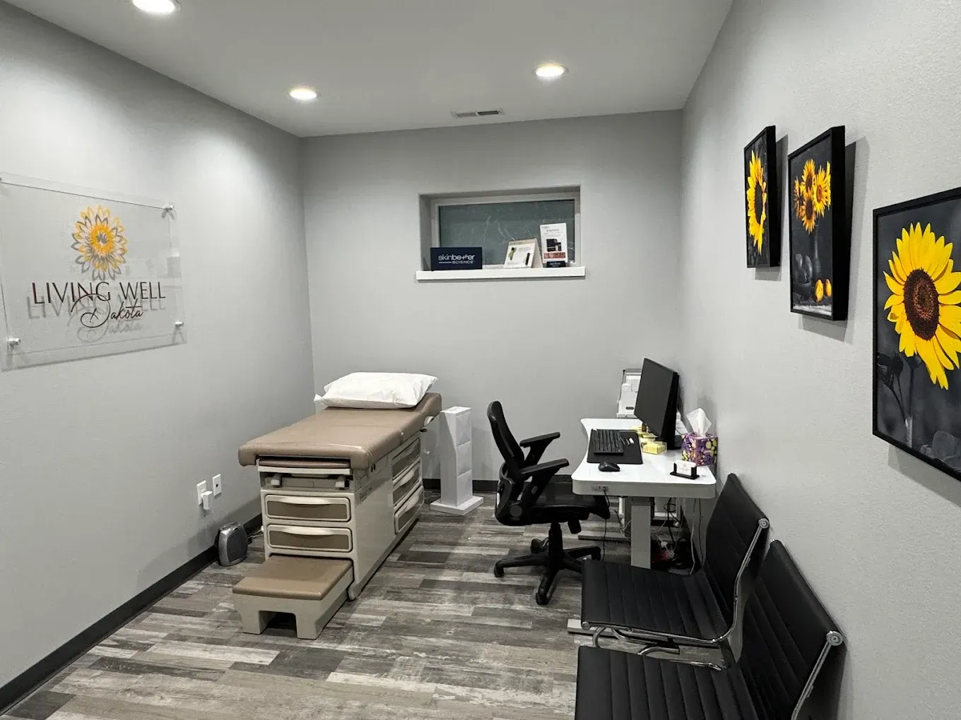 Small treatment room with massage table, desk, chairs, wall art, and sunflowers on gray walls