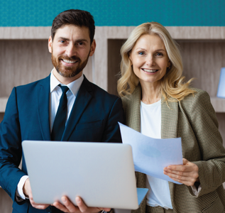 Two smiling professionals: a man with a beard in a suit holding a laptop, and a woman with papers, in an office setting.