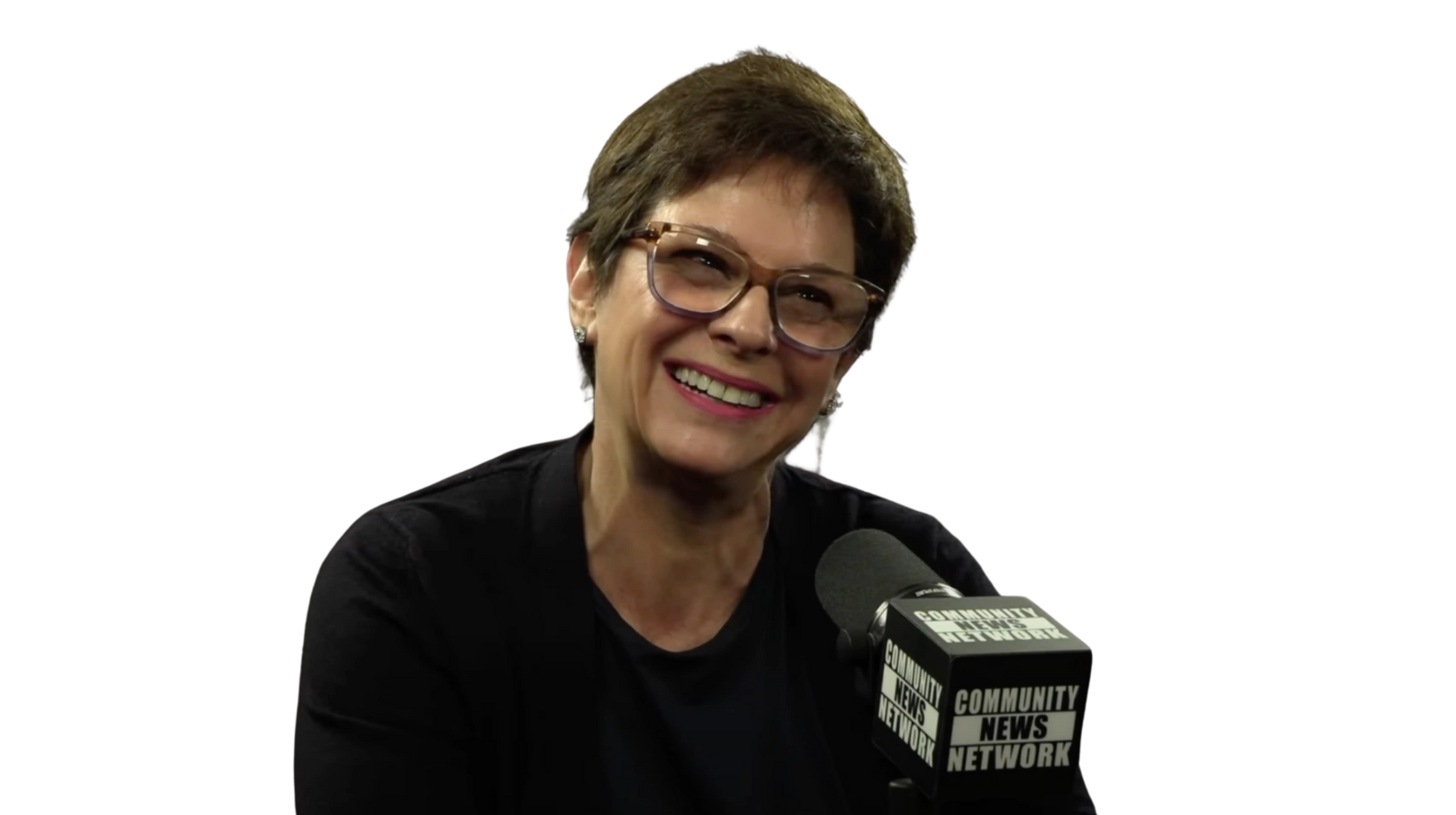 Man in black shirt and cap gesturing, seated near a microphone, white background.