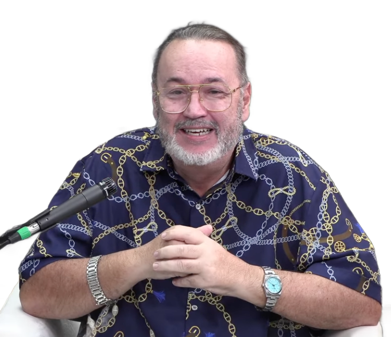 Man with glasses, gray beard, in blue shirt, smiling, sitting, hands clasped.