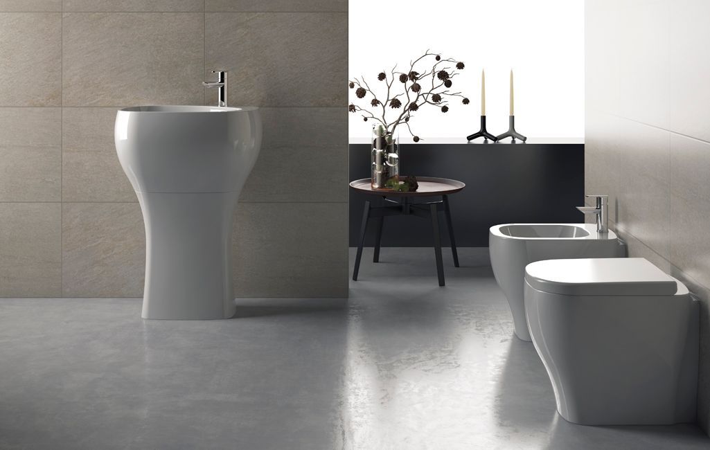 Bagno moderno con lavabo a colonna bianco, vasca da bagno e due WC su pavimento piastrellato grigio.