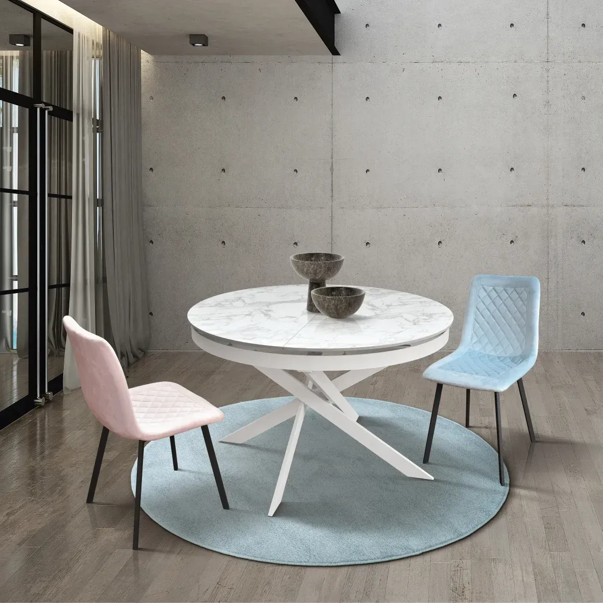 Angolo pranzo moderno con tavolo rotondo bianco, sedie rosa e blu e piccole ciotole su uno sfondo di cemento.