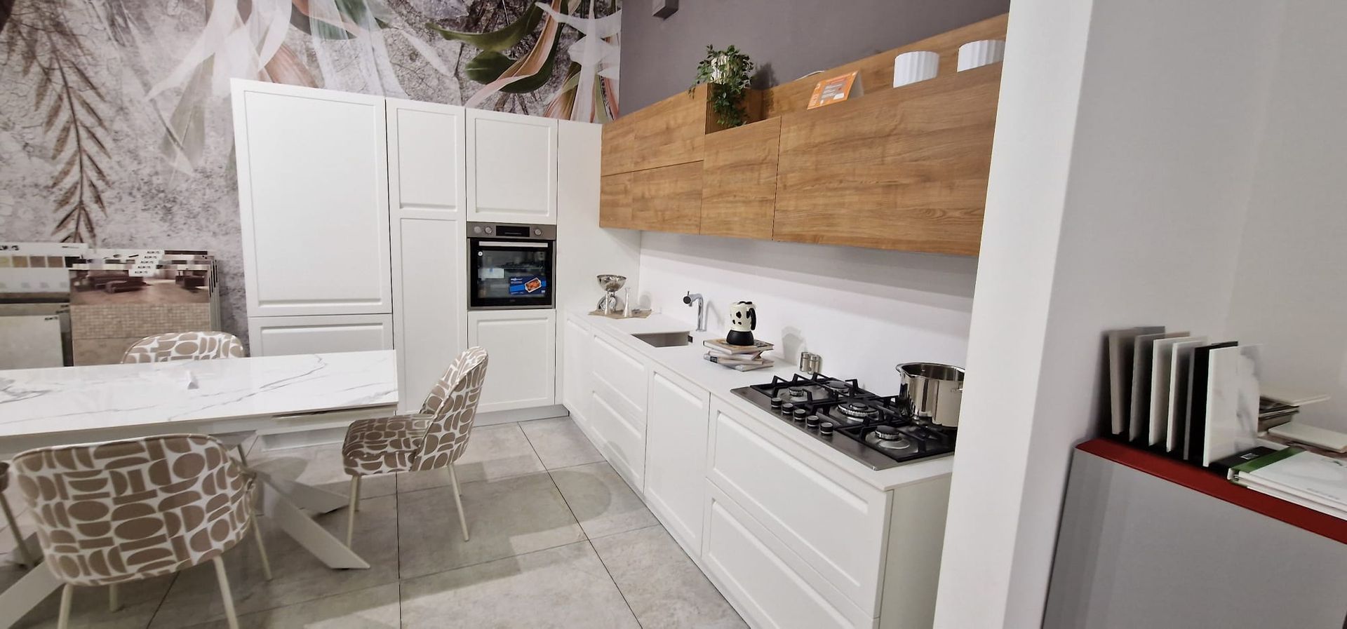 Cucina moderna bianca con zona pranzo, forno a incasso, paraschizzi in legno e tavolo vicino alla finestra.