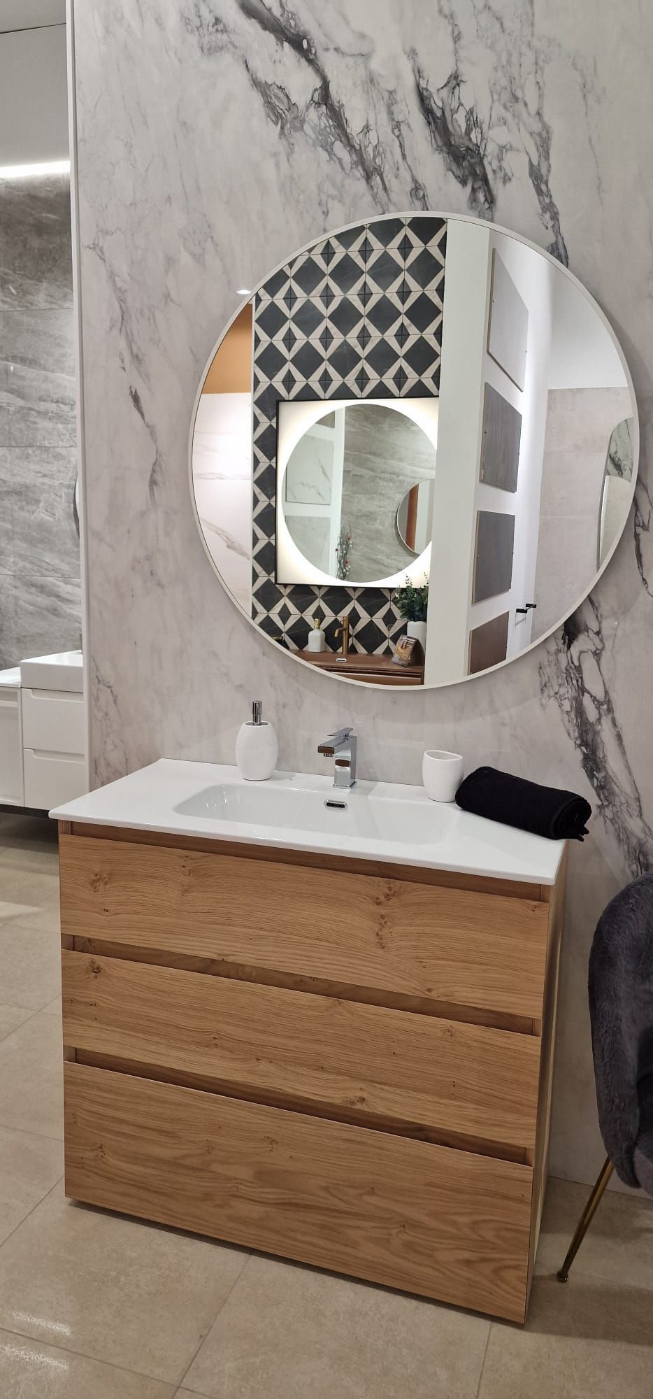 Mobile da bagno con cassetti in legno, lavabo bianco, specchio rotondo e parete con rivestimento effetto marmo.