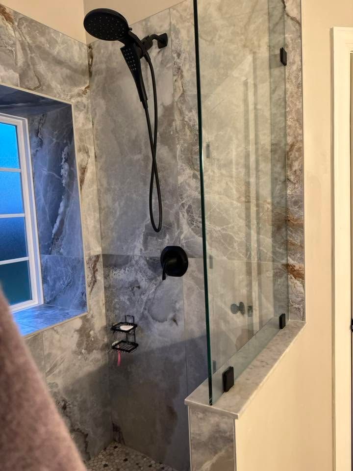 Walk-in shower with gray tile, black showerhead, and glass partition beside a small window
