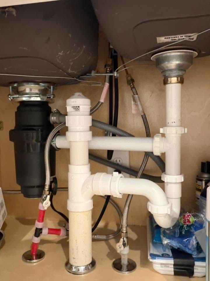 Under-sink plumbing with white PVC pipes and a black garbage disposal beneath a kitchen sink.