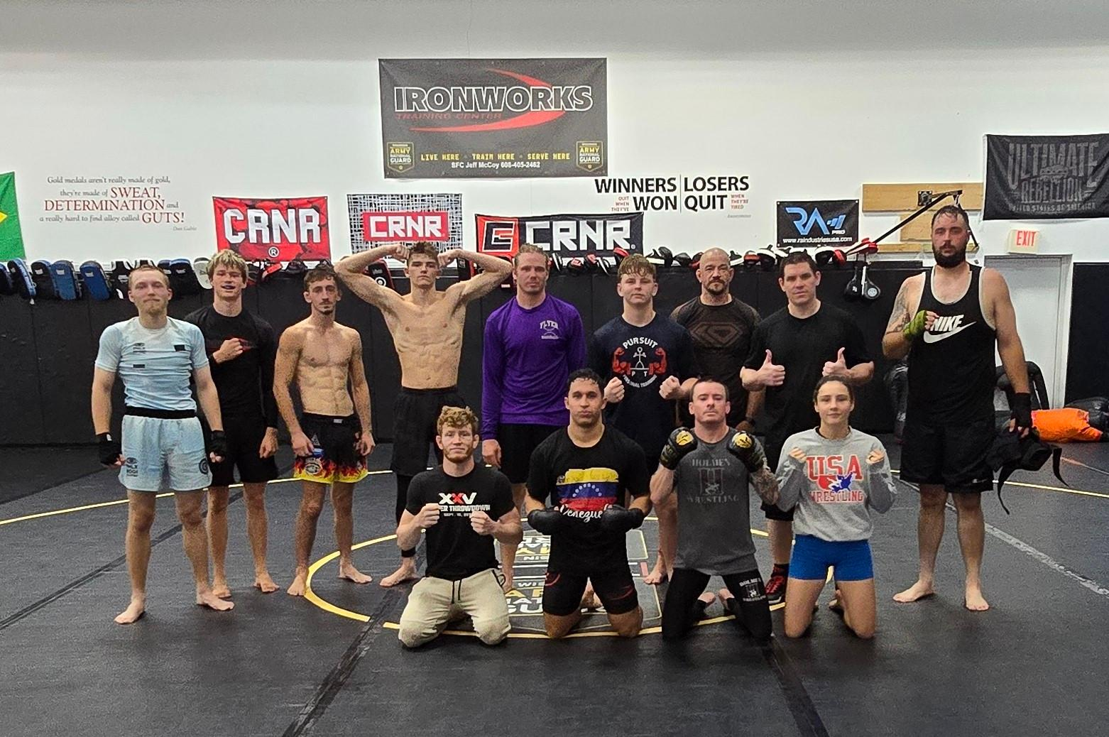 Group of athletes posing in a gym, some holding championship belts, with banners on the wall behind them.