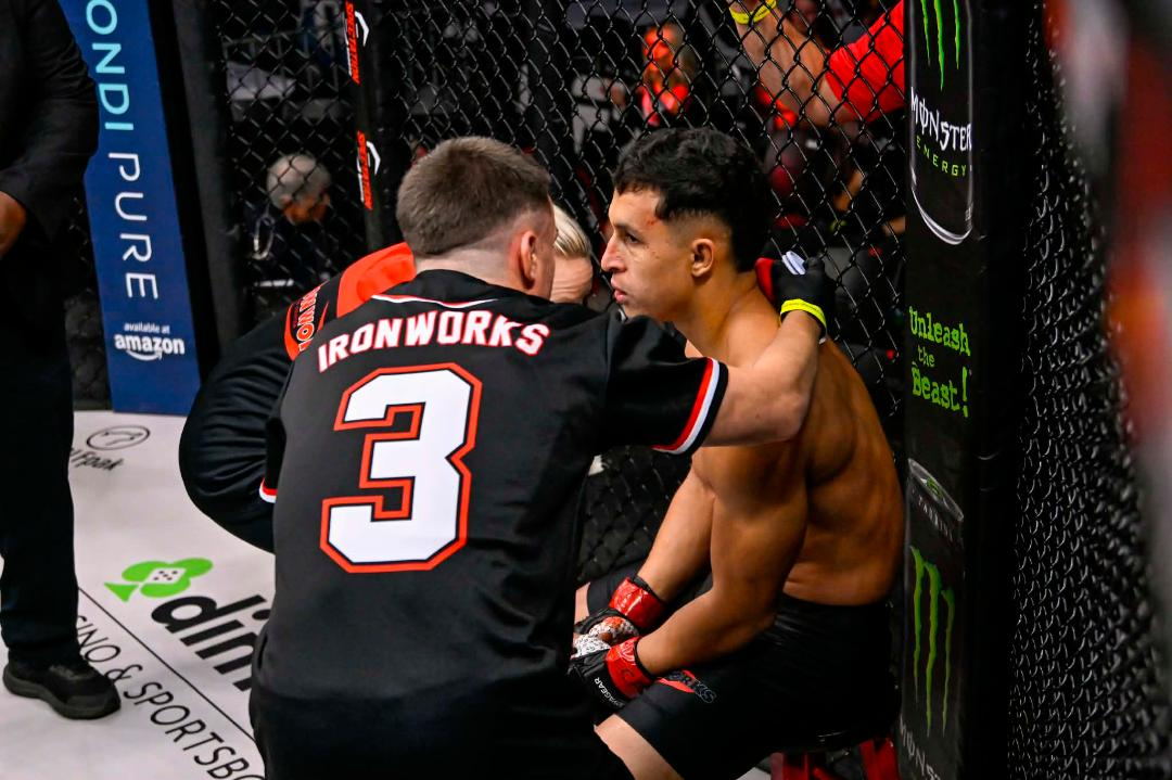 MMA corner coach in black jersey with red “3” tending to seated fighter inside the cage