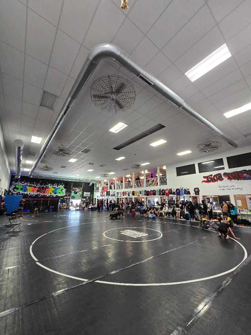 Indoor wrestling gym with a black mat, circular markings, and people seated around the perimeter