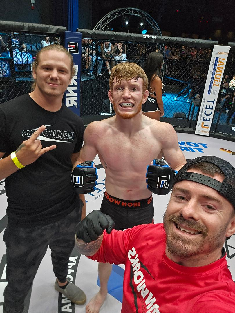 Three men posing in a cage arena, one shirtless with gloves, in a mixed martial arts setting.