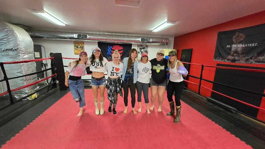Six people posing in a boxing ring with red flooring and walls, smiling toward the camera.