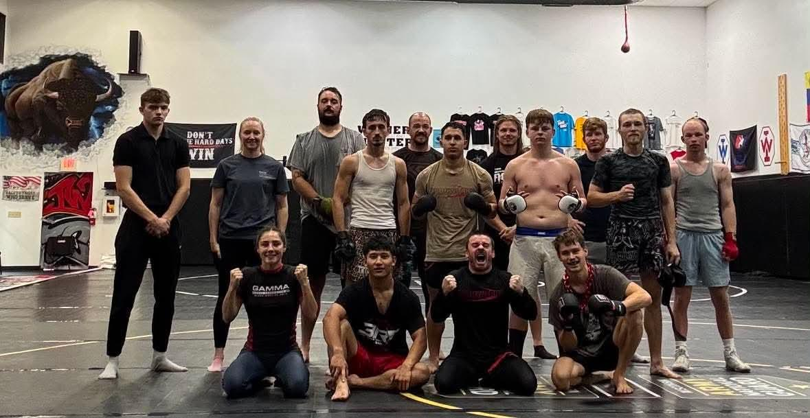 Group of people posing together in a gym, with kettlebells and workout equipment in the background.