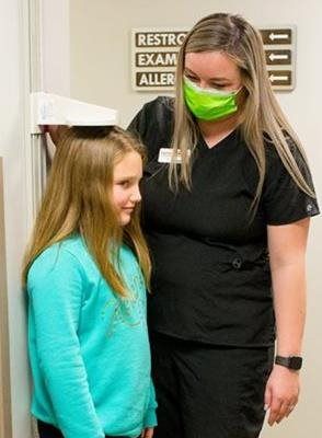 Girl's height being measured by a person in a medical office, both wearing masks.