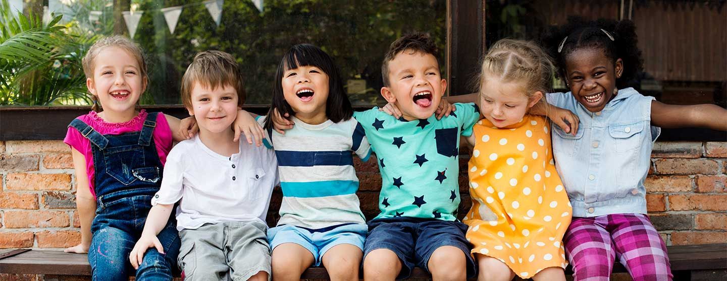 A group of smiling children with arms around each other sitting on a bench outside.