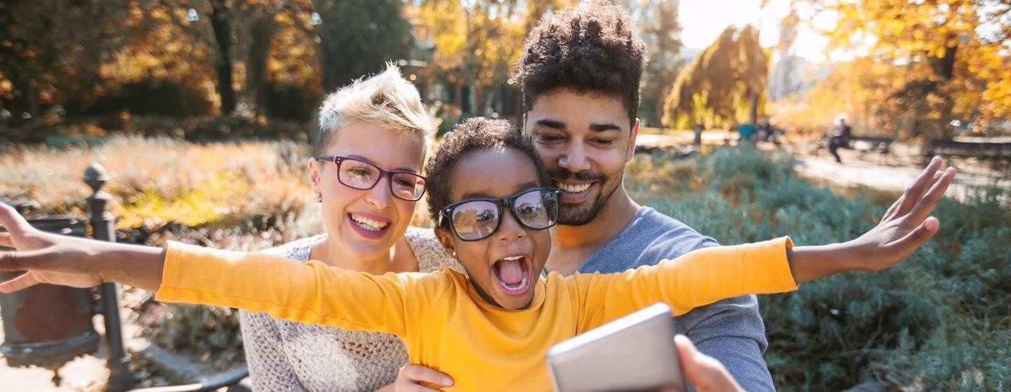 Three friends smiling outdoors, one holding a tablet and another spreading arms wide in sunlight