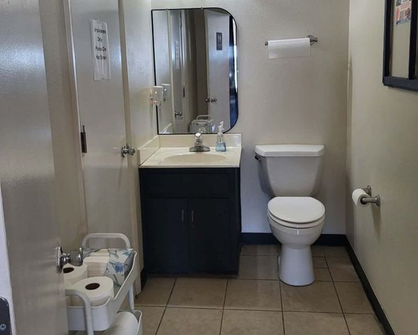 Bathroom with sink, toilet, mirror, and a small cart with paper products. Beige tile floor.