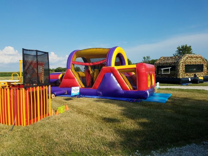 Inflatable obstacle course with red, purple, and yellow sections on a grassy field; dunk tank is nearby.