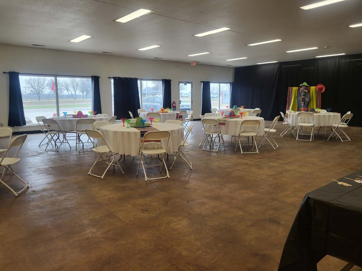 Large event room with white-clothed tables, chairs, and decor near windows. Brown floor, dark curtains, and overhead lights.
